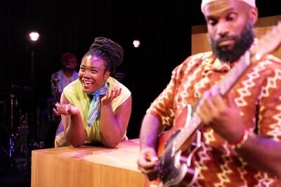 Scène uit De Suriname-trilogie: Stepping Stones: een speelster met opgestoken vlechten leunt lachend op een houten blok en gebaart met beide handen. Op de voorgrond staat onscherp een gitarist in een bruin gebatikt hemd.