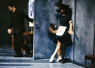 Beeld bij Out of the stillness: een danseres in een dienstbodejurk met wit schort wordt tegen een metalen deur geworpen, haar haren in de lucht en een zilveren dienblad in haar hand. Links loopt een man in een donker pak weg.