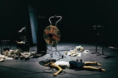 Beeld bij Out of the stillness: een danser ligt languit op de vloer van een donker podium, naast een grote staande ventilator. Om hem heen liggen verfrommelde papieren, kabels en theaterlampen.