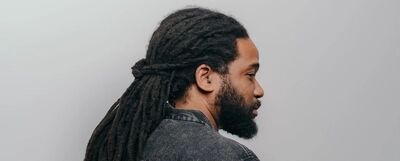 Campagnebeeld voor Malebox: een man met lange dreadlocks in een staart en een volle baard staat in profiel voor een grijze achtergrond, in een spijkerjasje.