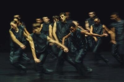 Een groep dansers in Everywhere is here beweegt zijwaarts over een donker toneel; de lange sluitertijd maakt de bewegingen tot vegen, één danser in het midden staat scherp.