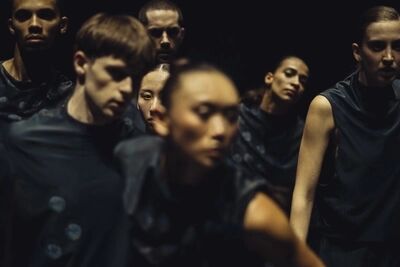 Beeld bij Everywhere is here: een groep dansers in donkerblauwgrijze shirts met lichte ronde motieven staat dicht opeen in het donker. De gezichten kijken verschillende kanten op; de danser vooraan is onscherp.