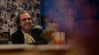 Campagnebeeld voor Wachten met Dirk De Wachter: een man met halflang grijzend haar, een zwart colbert en een lichte sjaal zit lachend aan een tafel in een café. Achter hem hangt een wand vol posters en foto's; op de voorgrond staat onscherp een waxinelichtje.