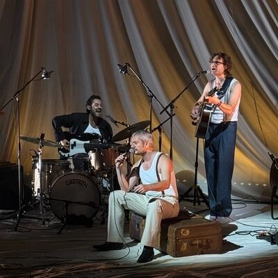In Wachtend op de dood zingt een man met grijs haar en een snor, gezeten op een oude koffer, in een microfoon, in een wit hemd met bretels; links speelt een man achter een drumstel met 'GRETSCH' basgitaar en rechts een man met een bril gitaar, beiden zingend, voor plooiende lichte doeken.