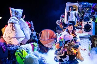 Scène uit Waste me (13+): drie spelers dragen kostuums van huisraad — links iemand gewikkeld in dekbedden met een kussen om het hoofd, rechtsboven iemand met een matras om het lichaam en vooraan een speler behangen met knuffelbeesten. Achter hen staan een wasmachine, een tent en stapels spullen.